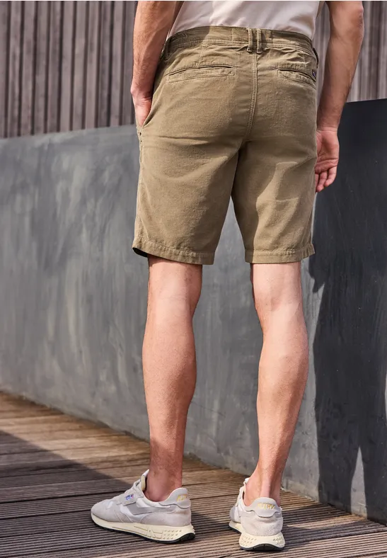 Ein Mann steht auf einer Holzterrasse, trägt khakifarbene Shorts und weiße Sneaker, Rückenansicht.