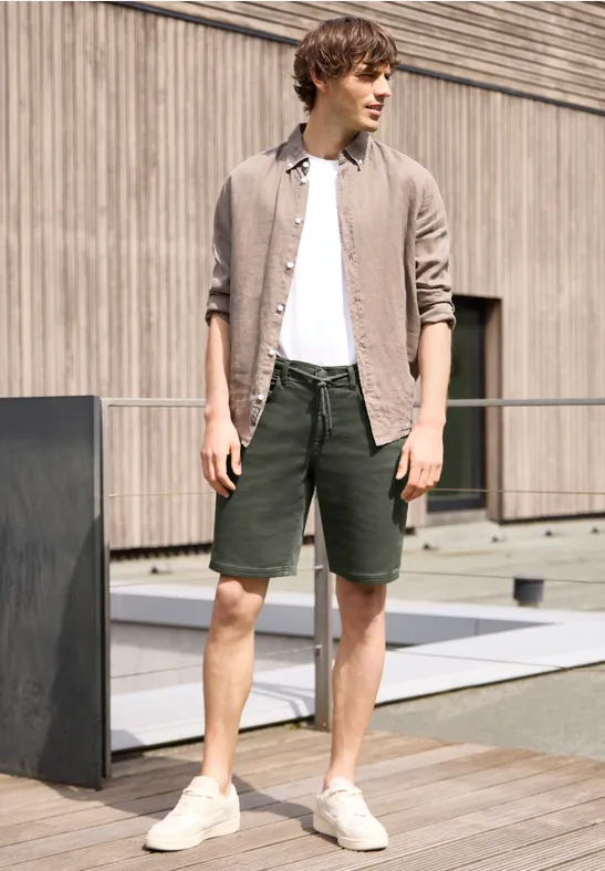 Homme en T-shirt blanc, chemise beige ouverte, short vert et baskets blanches devant un mur en bois.