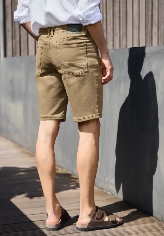 Ein Mann mit beiger Short, weißem Hemd und Sandalen steht seitlich vor einer Holzwand im Sonnenlicht.