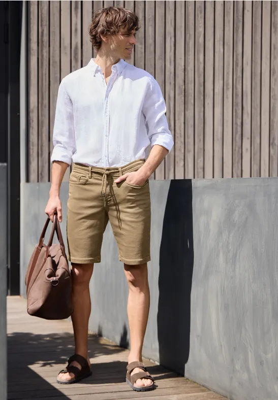 Ein Mann in weißem Hemd und braunen Shorts trägt Tasche, steht neben Holzfassade.