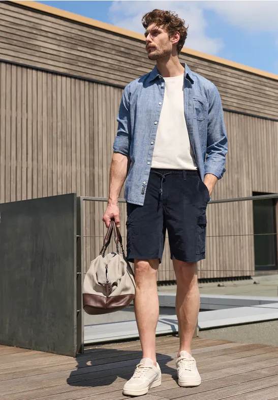Ein Mann in Hemd, T-Shirt, Shorts und Sneakern steht draußen und hält eine Reisetasche.