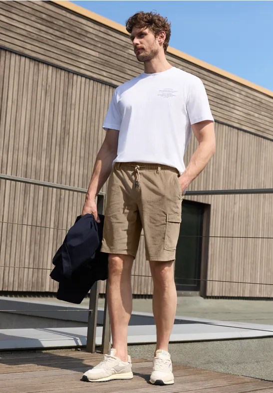 Mann mit weißem T-Shirt und khakifarbenen Cargo-Shorts, hält Jacke, vor modernem Gebäude.