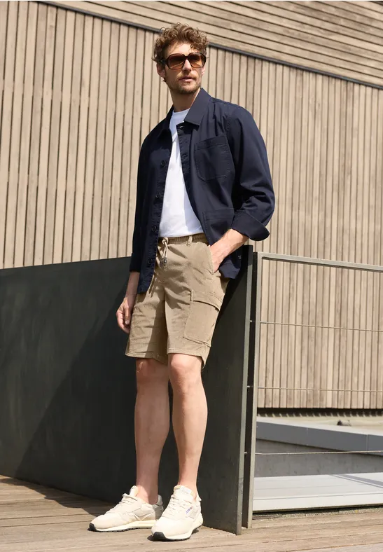 Ein Mann in beigen Shorts, weißem T-Shirt und dunkelblauer Hemdjacke lehnt an einer Holzfassade.