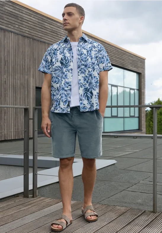 Mann auf Dachterrasse vor Holzglasgebäude, trägt blau-weißes Hemd, weißes Shirt, graue Shorts und Sandalen.