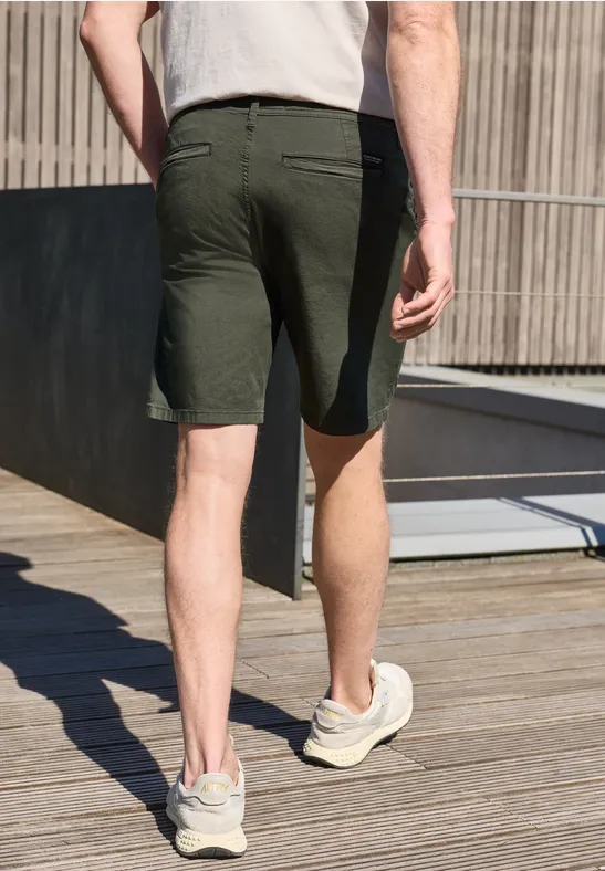 Man van achteren in donkergroene short en sneakers, lopend op houten terras bij gebouw.