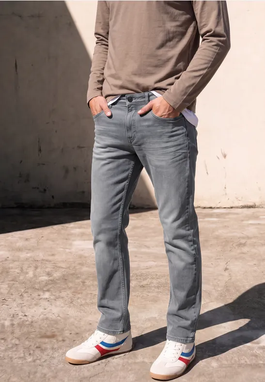 Mężczyzna w szarych dżinsach, longsleeve i białych sneakersach z czerwono-niebieskimi paskami przy betonowej ścianie.