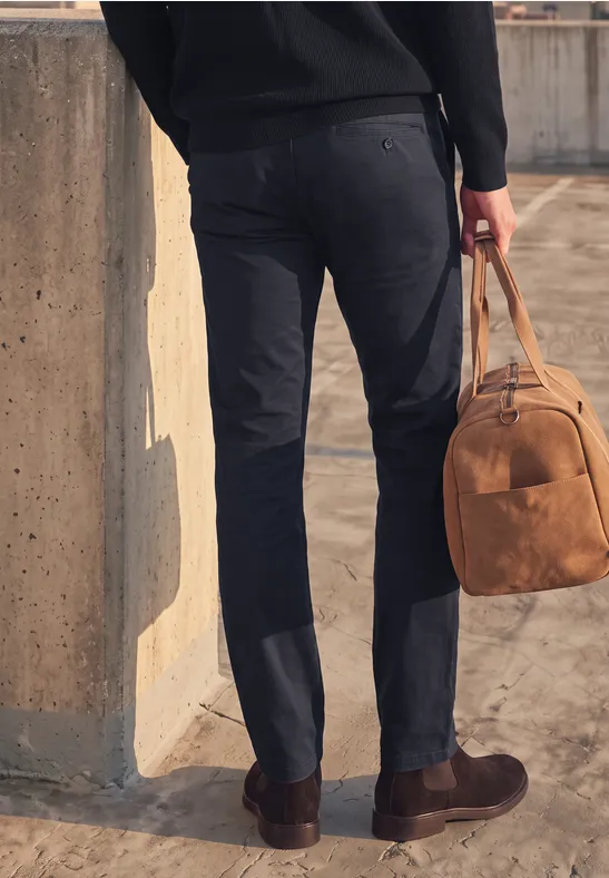 Eine Person steht neben einer Betonwand, trägt dunkle Hose und Pullover und hält eine braune Tasche.