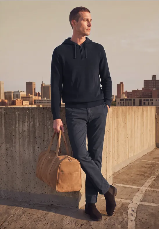 Mann steht auf Dach mit Stadtblick, trägt lässiges Outfit und hält eine braune Reisetasche in der Hand.