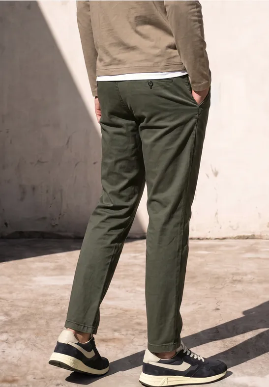 Rückansicht einer Person in dunkelgrünen Chinos und schwarzen Sneakers, stehend im Sonnenlicht.