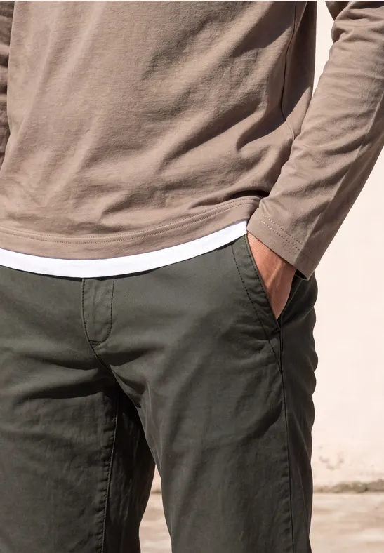 Person in hellbraunem Langarmshirt über weißem T-Shirt und dunkelgrüner Hose, Hand in Tasche
