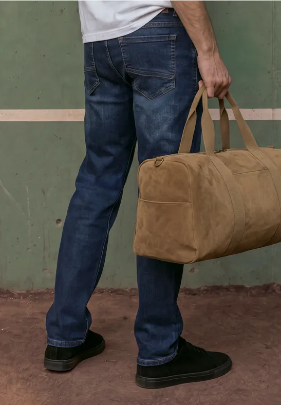 Mann trägt blaue Jeans und schwarze Schuhe, hält eine beige Reisetasche vor einer grünen Wand.