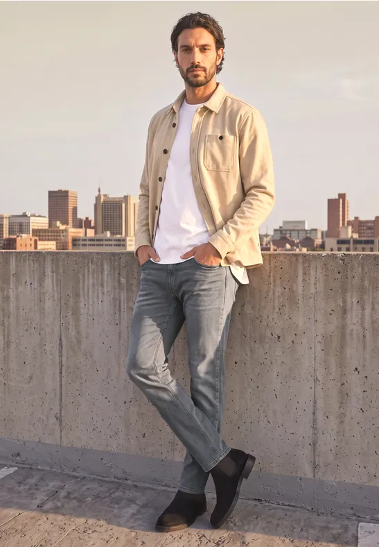 Mann mit Jeans und beiger Jacke lehnt an Betonmauer, Stadtsilhouette im Hintergrund.