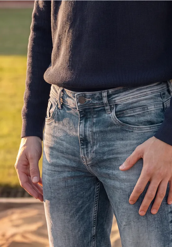 Eine Person trägt draußen blaue Jeans und einen dunklen Pullover und steht auf sandigem Untergrund.