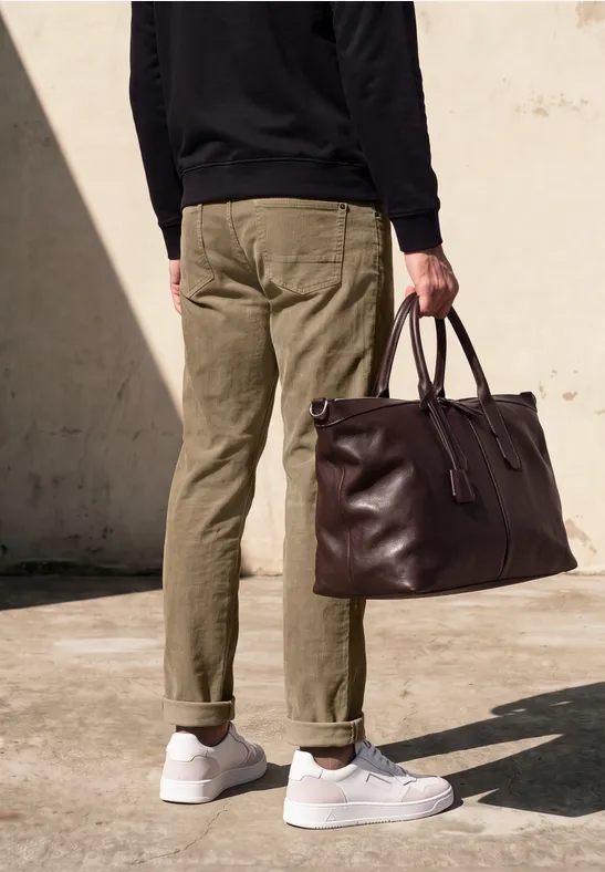 Person in khakifarbenen Hosen und weißen Sneakern hält große braune Lederreisetasche.