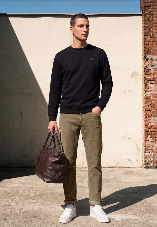Ein Mann in schwarzem Pullover, grünen Hosen und weißen Sneakern trägt braune Tasche.