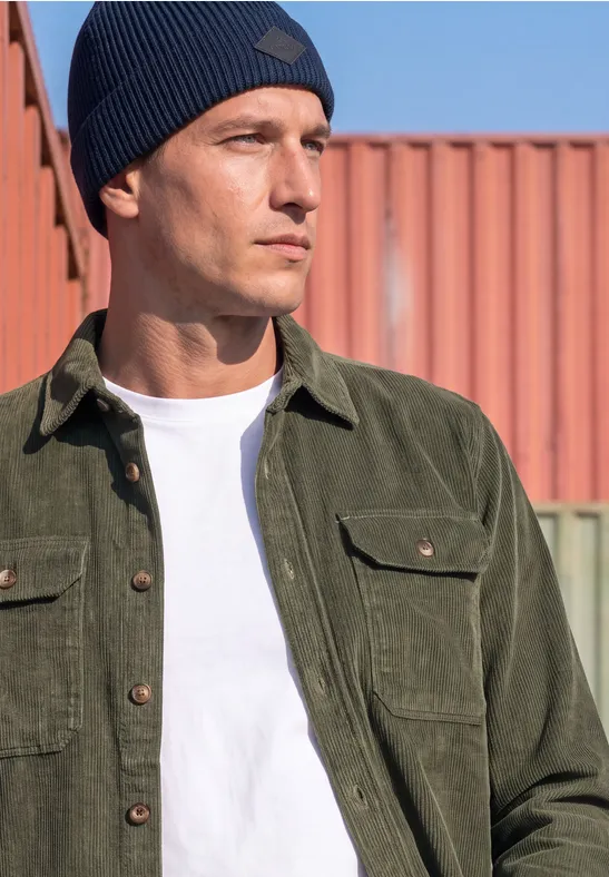 Ein Mann in weißem T-Shirt, grüner Cordjacke und blauer Mütze blickt seitlich im urbanen Umfeld.