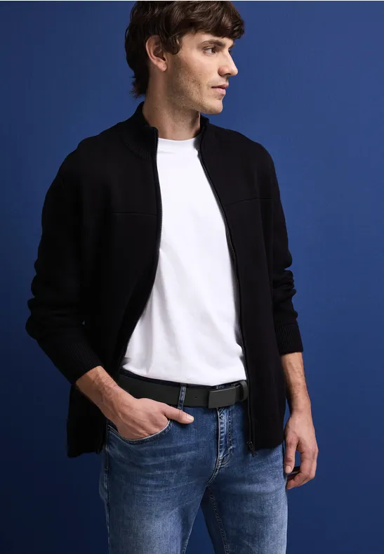 Mann mit weißem T-Shirt, schwarzer Jacke und Jeans vor blauem Hintergrund.