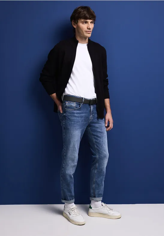 Mann in Jeans, weißem T-Shirt und schwarzer Jacke steht vor blauer Wand.