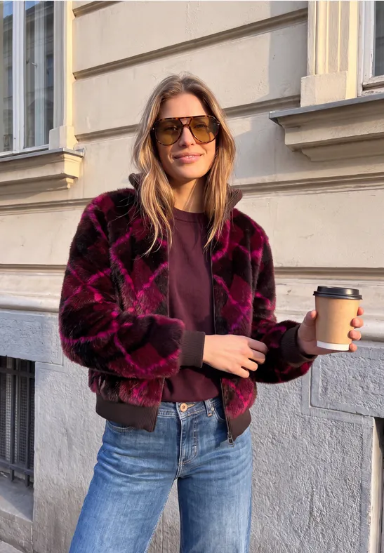 Frau in stylischer Jacke hält To-go-Kaffeebecher vor urbaner Fassadenwand