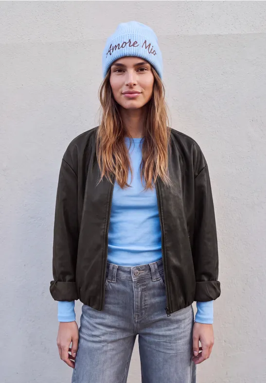 Junge Frau mit blauer Mütze, hellblauem Shirt, grauen Jeans und dunkler Jacke vor hellem Hintergrund.