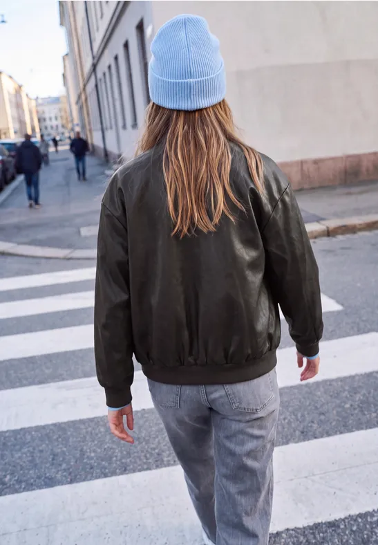 Eine Person mit blauer Mütze und grüner Jacke geht über einen Zebrastreifen in einer Stadt.