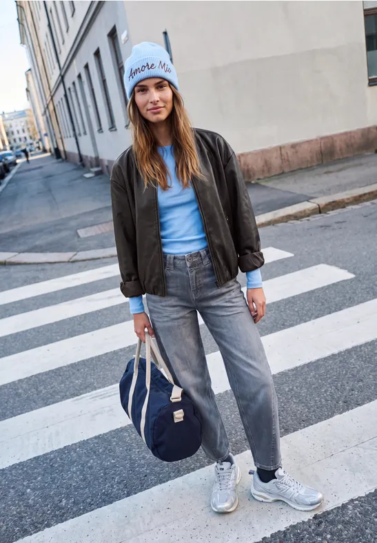 Junge Frau in Jeans, Bomberjacke und Mütze mit Tasche steht auf einem Zebrastreifen.