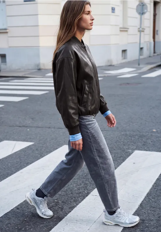 Junge Frau in Lederjacke, Jeans und Sneakers läuft über einen Zebrastreifen in der Stadt.