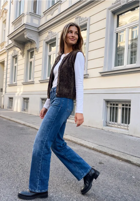 Junge Frau in Jeans, weißem Shirt und brauner Weste geht lächelnd an elegantem Stadthaus vorbei.