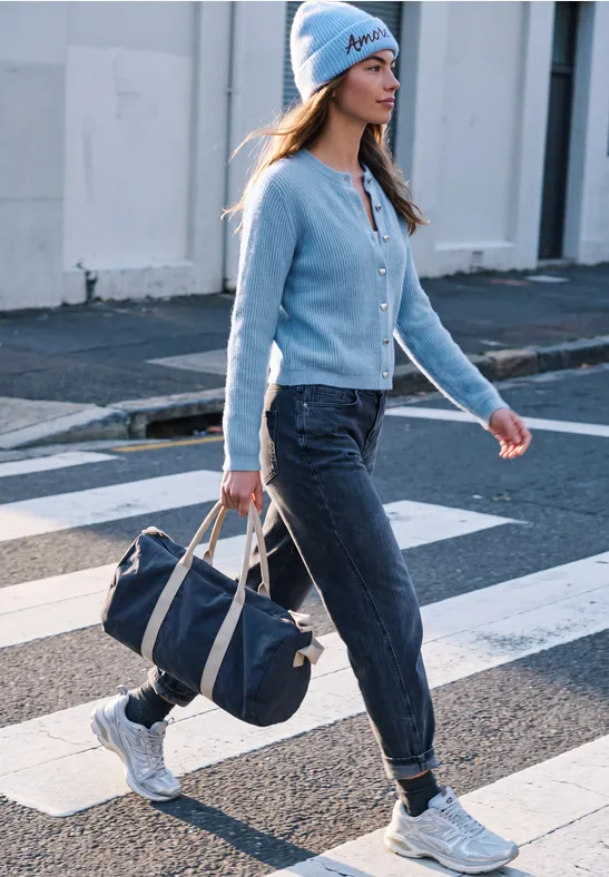 Eine Frau überquert einen Zebrastreifen, trägt blaue Mütze, Jacke, dunkle Hose und weiße Sneaker, hält Tasche.