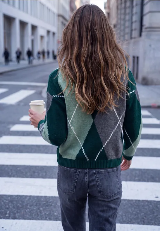 Eine Person in grauen Jeans und grün gemustertem Pullover mit Kaffee in der Hand, die eine Straße überquert.