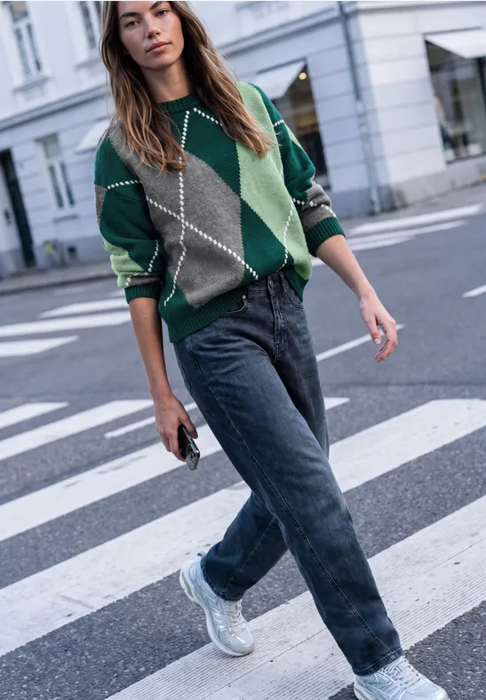 Frau in grün-grauem Patchwork-Pullover, Jeans und weißen Sneakern überquert eine Straße und hält ein Handy.