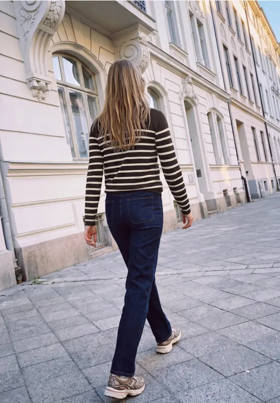 Eine Person mit gestreiftem Pullover und Jeans geht auf einem Bürgersteig an Gebäuden vorbei.