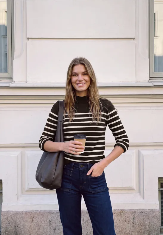 Eine junge Frau steht vor einer Fassade, hält einen Coffee-to-go und trägt Ringelshirt und Jeans.