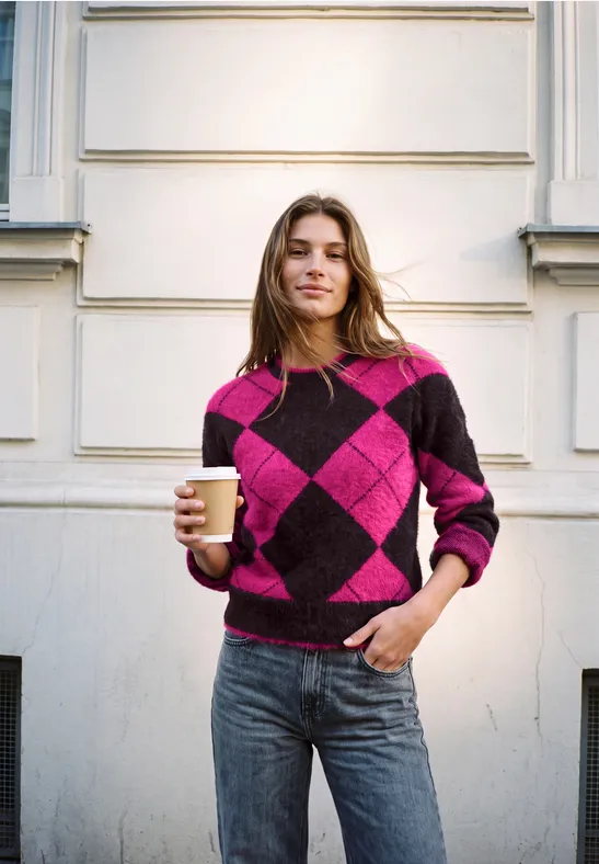 Junge Frau im pink-lila-Rautenpullover hält einen Coffee-to-go-Becher vor einer weiß-gestrichenen Hauswand.