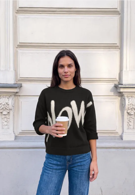 Frau mit LOVE-Pullover hält einen Kaffee vor einer hellen Hausfassade.