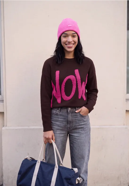 Femme souriante portant un bonnet rose, un pull marron "NOW", un jean et tenant un sac bleu devant un mur neutre.