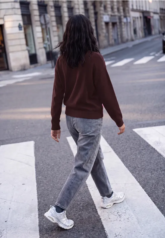 Femme en veste brune et jeans gris traverse un passage piéton en ville