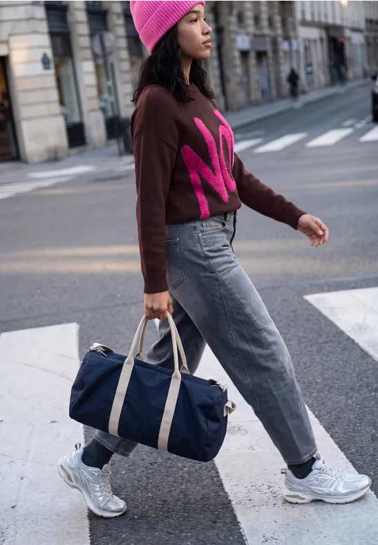 Femme en bonnet rose et pull traverse la rue en ville, portant un sac de voyage bleu et des baskets.
