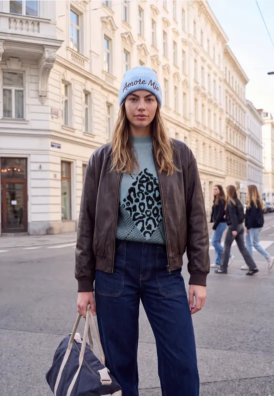 Jonge vrouw in jeans, leren jas en luipaardtrui staand in een stedelijke straat.