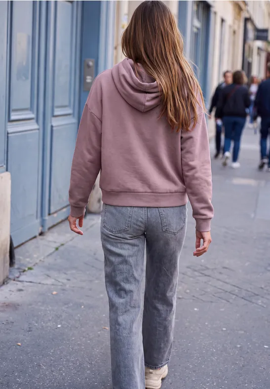 Eine Person in rosa Kapuzenpullover und grauen Hosen geht auf einem Bürgersteig in der Stadt.
