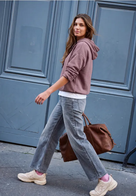 Eine junge Frau in Hoodie und Jeans trägt eine große Tasche und geht an einer blauen Tür vorbei.