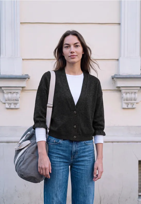 Jeune femme en cardigan, t-shirt blanc, jean et sac, debout devant une façade claire.