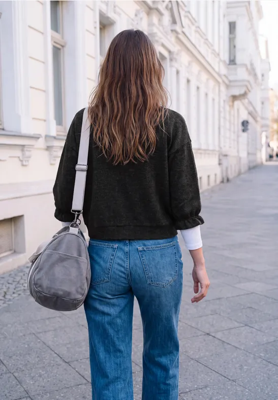 Une femme en jean bleu et pull noir marche dans la rue, portant un sac gris.