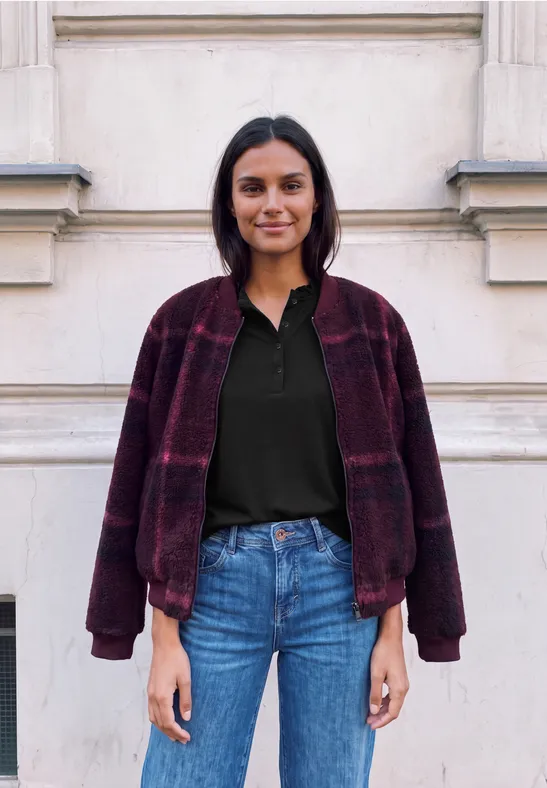 Eine Frau in Jeans, schwarzem Shirt und weinrotem Karomantel steht vor einer hellen Hauswand.