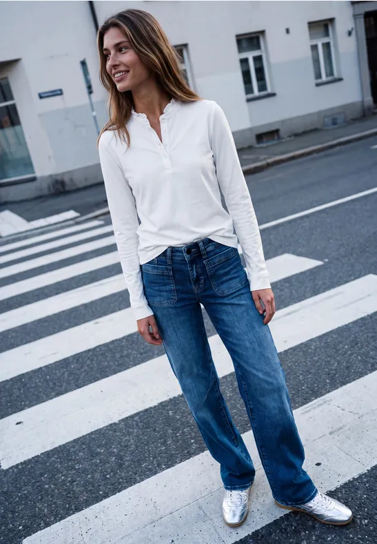 Frau in weißer Bluse und Jeans steht auf einem Zebrastreifen in urbaner Umgebung.