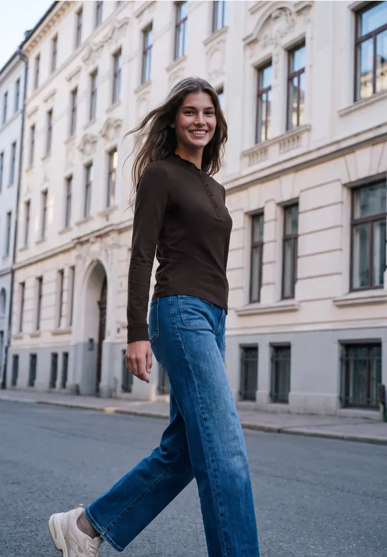 Frau in Jeans und dunklem Pullover lächelt entspannt beim Spaziergang auf einer ruhigen Stadtstraße.