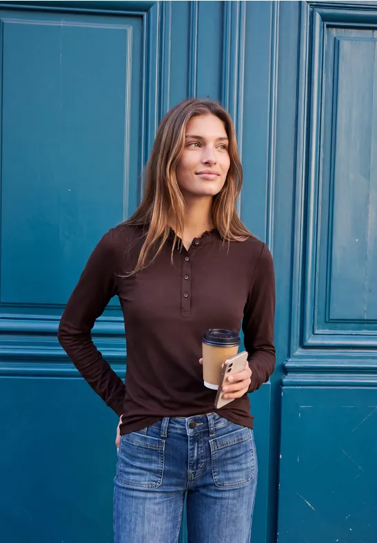 Junge Frau lehnt vor türkisem Türflügel, trägt braunes Longsleeve, Jeans und hält Kaffeebecher, lächelt.