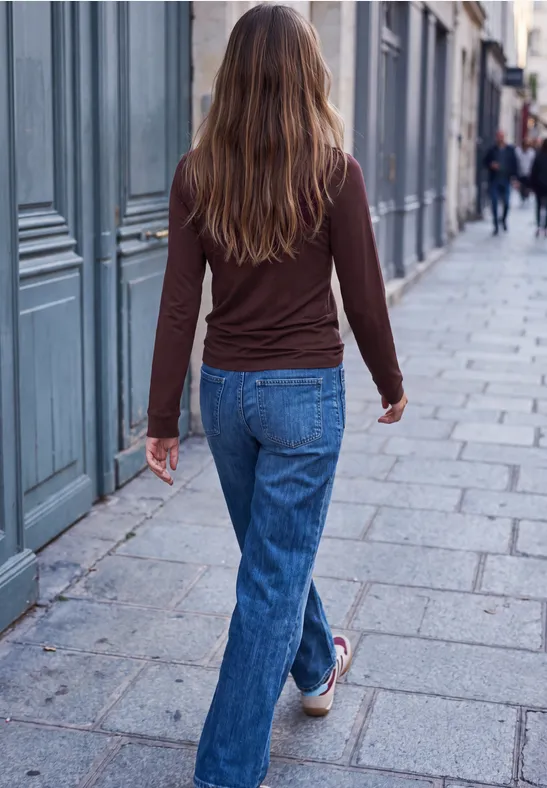 Eine Frau in Jeans und braunem Oberteil geht eine gepflasterte Straße entlang, aufgenommen von hinten.