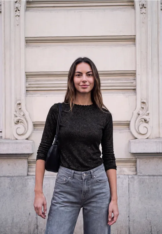 Eine Frau in schwarzem Pullover und Jeans steht vor einer hellen Fassade und blickt in die Kamera.