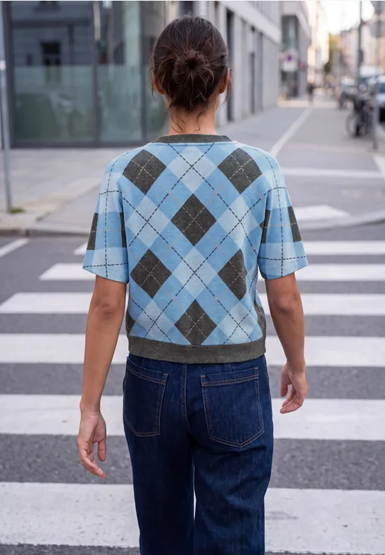 Frau in blauem Rautenmuster-Pullover und Jeans steht auf einem Zebrastreifen in der Stadt.